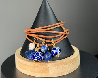 Pode incluir: Uma pulseira de enrolar com cordão de couro castanho. A pulseira apresenta berloques dourados com contas estampadas em azul e branco, uma pérola e uma borla azul marinho. A pulseira é exibida em um suporte cônico preto.