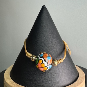 May include: A handmade bracelet featuring a large, colorful bead with orange, blue, yellow, and green accents. The bead is strung on a gold-colored cord with gold-toned metal beads on either side. The bracelet is displayed on a black cone-shaped stand.