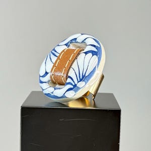 May include: A unique ring featuring a white and blue ceramic oval design with a brown leather band in the center. The ring has a gold-colored metal band. The ceramic design has a floral pattern.