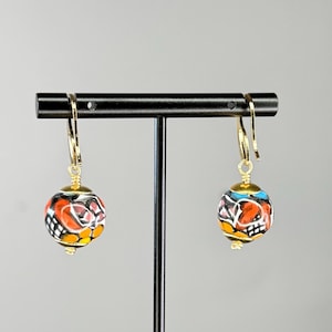 May include: A pair of handcrafted, colorful beaded earrings. Each earring features a round, painted bead with a gold-colored hook. The beads display a mix of orange, red, yellow, black, and blue patterns, creating a unique and artistic design.