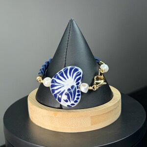 Pode incluir: Pulseira em forma de cora&ccedil;&atilde;o azul e branco com detalhes dourados e p&eacute;rolas falsas. A pulseira apresenta um cord&atilde;o azul e um pingente decorativo em forma de cora&ccedil;&atilde;o com um padr&atilde;o detalhado em azul e branco. &Eacute; exibida em um suporte preto.