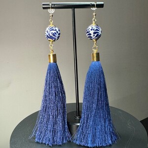 May include: A pair of long, dangling earrings. Each earring features a navy blue tassel, a gold-colored metal cap, a white and blue patterned bead, and a gold-colored wire hook. The earrings are displayed on a black stand.