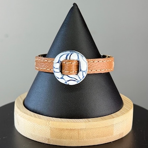 May include: A brown leather bracelet with a white and blue patterned circular buckle. The bracelet is displayed on a black cone-shaped stand with a wooden base. The leather band is stitched with a light brown thread.