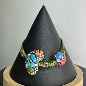 May include: A colorful bracelet with a green cord and two decorative ceramic pieces. One piece is heart-shaped with blue flowers, and the other is a round bead with a mosaic design. The bracelet is displayed on a black stand.