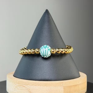 May include: A beaded ring featuring a series of small, polished gold-tone metallic spheres arranged in a circular band. A single, larger round bead with a white and teal green botanical or leaf-like pattern is centered on the front of the ring.