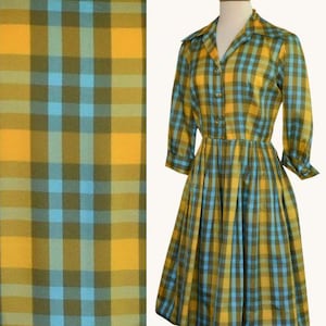 May include: A vintage-style dress in a yellow, blue, and olive green plaid pattern. The dress features a collared neckline, button-down front, and a flared skirt. The sleeves are three-quarter length, and the dress is displayed on a mannequin.