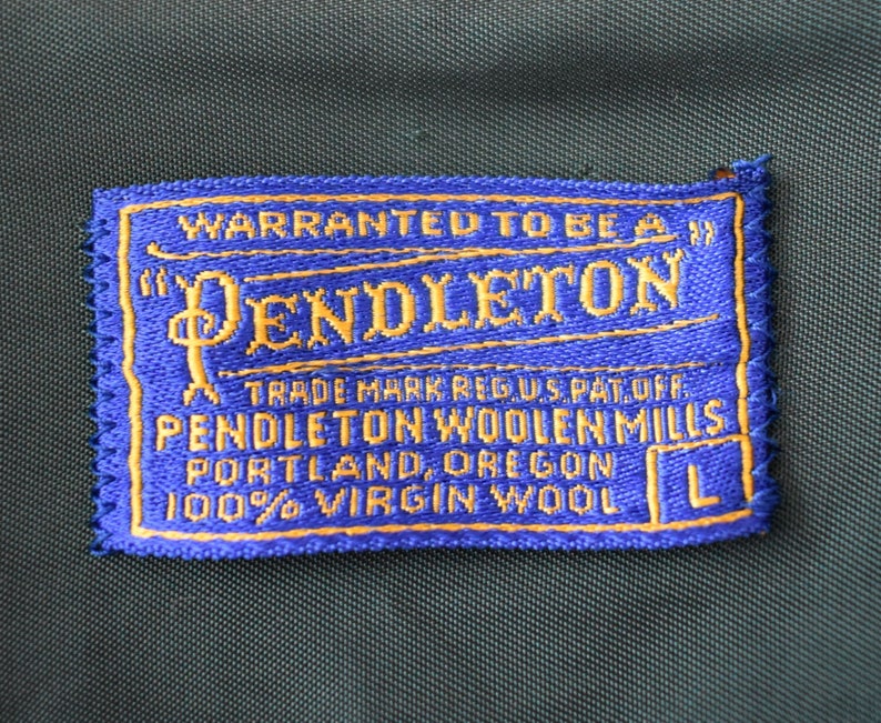 Puede incluir: Una etiqueta Pendleton azul y dorada con el texto "WARRANTED TO BE A" y "PENDLETON". La etiqueta tambi&eacute;n incluye "TRADE MARK REG US PAT OFF", "PENDLETON WOOLEN MILLS", "PORTLAND, OREGON" y "100% VIRGIN WOOL".
