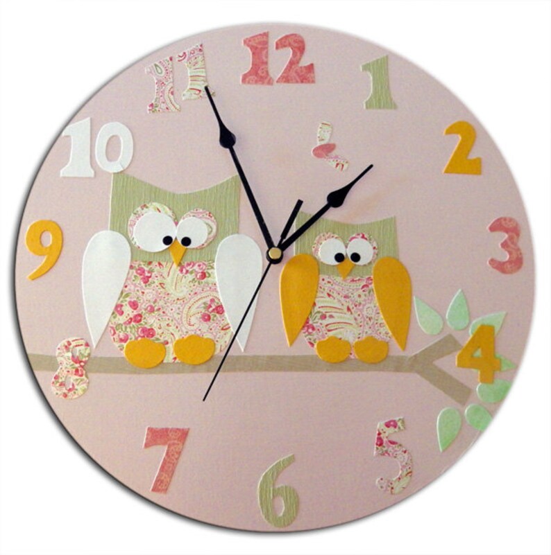 Puede incluir: Reloj redondo rosa con dos b&uacute;hos, uno a cada lado de una rama. Los b&uacute;hos est&aacute;n hechos de tela con un patr&oacute;n floral y tienen picos y patas amarillas. La esfera del reloj tiene n&uacute;meros blancos del 1 al 12 y manecillas negras.