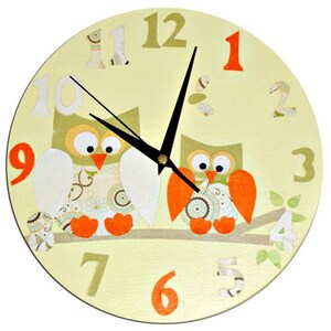 Puede incluir: Un reloj redondo amarillo con dos b&uacute;hos posados en una rama. Los b&uacute;hos son verdes y marrones con patas naranjas y tienen ojos grandes. La esfera del reloj tiene agujas negras y n&uacute;meros naranjas.