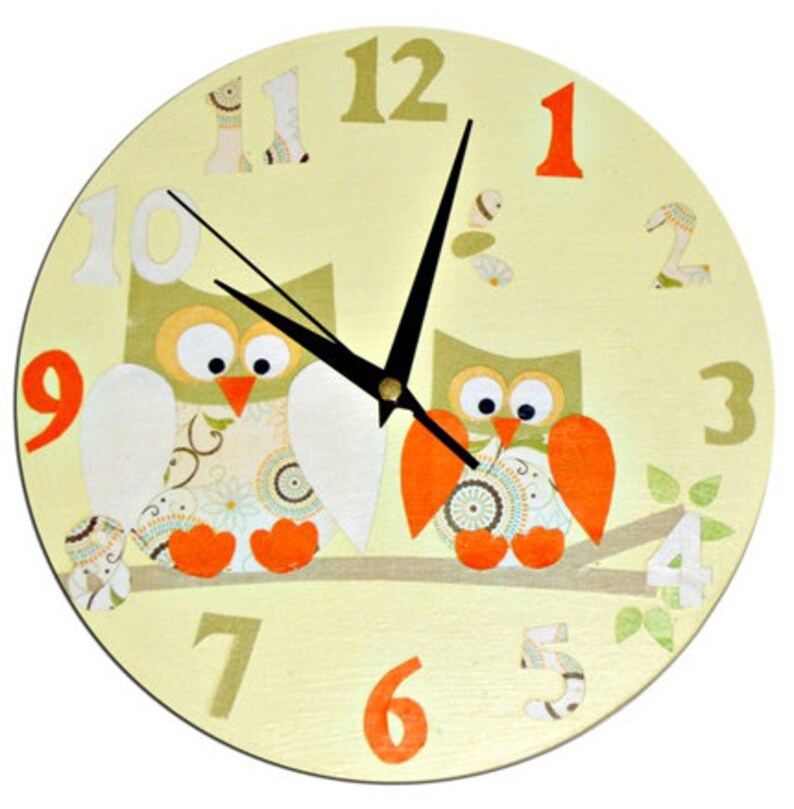 Puede incluir: Un reloj redondo amarillo con dos b&uacute;hos posados en una rama. Los b&uacute;hos son verdes y marrones con patas naranjas y tienen ojos grandes. La esfera del reloj tiene agujas negras y n&uacute;meros naranjas.