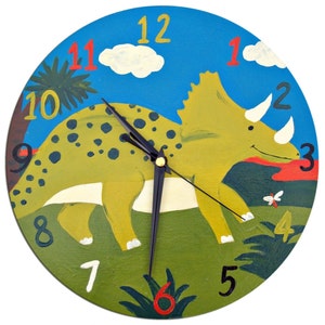 May include: A colorful wooden clock featuring a green, yellow, and brown dinosaur with black spots. The clock face has a blue background with white clouds and green leaves. The numbers 1 through 12 are printed in red and black around the clock face.