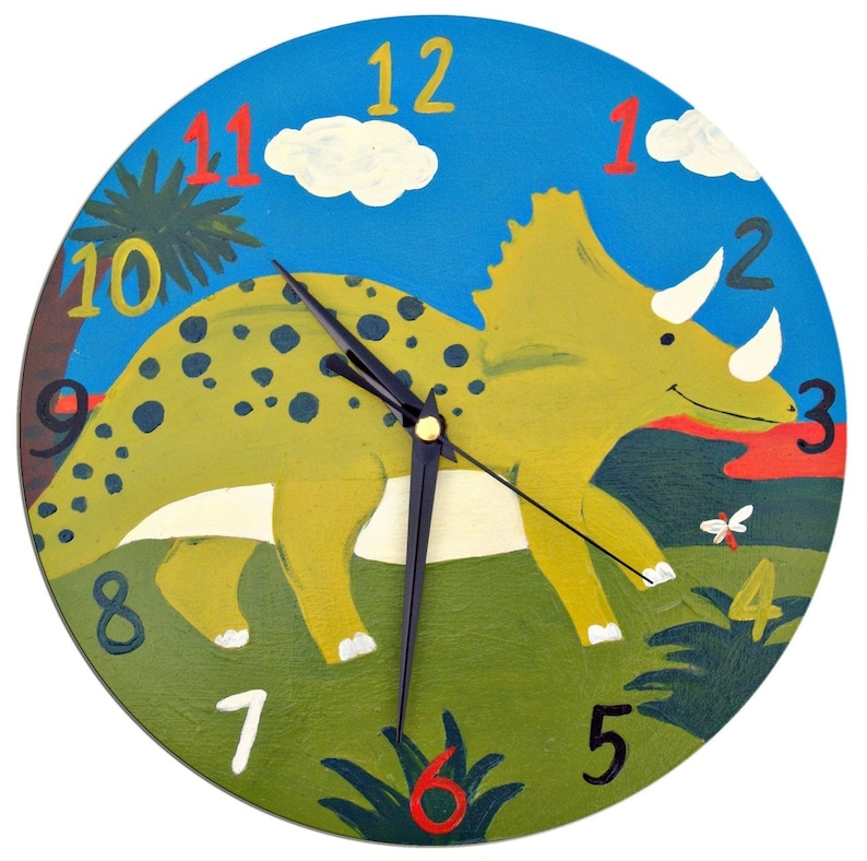 May include: A colorful wooden clock featuring a green, yellow, and brown dinosaur with black spots. The clock face has a blue background with white clouds and green leaves. The numbers 1 through 12 are printed in red and black around the clock face.