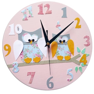 Puede incluir: Reloj redondo rosa con dos b&uacute;hos posados en una rama. Los b&uacute;hos son grises con alas de estampado floral y patas amarillas. La esfera del reloj tiene agujas negras y n&uacute;meros del 1 al 12 en varios colores y estilos.