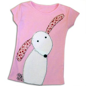 Op de afbeelding: Een roze T-shirt met een witte hond-applicatie. De hond heeft zwarte ogen en een zwarte neus. De oren van de hond zijn gemaakt van roze stof met een bloemenpatroon.