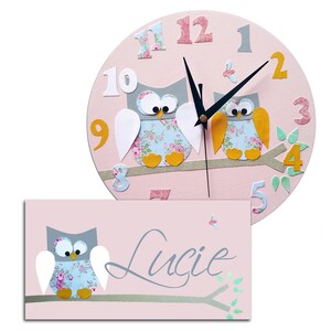 Puede incluir: Reloj de madera rosa en forma de b&uacute;ho con manecillas negras y una placa de nombre con el nombre "Lucie" en escritura cursiva. El reloj presenta dos b&uacute;hos con alas de estampado floral y patas amarillas, posados en una rama con hojas verdes.