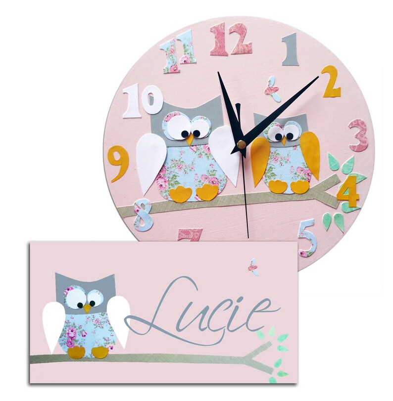 Puede incluir: Reloj de madera rosa en forma de b&uacute;ho con manecillas negras y una placa de nombre con el nombre "Lucie" en escritura cursiva. El reloj presenta dos b&uacute;hos con alas de estampado floral y patas amarillas, posados en una rama con hojas verdes.