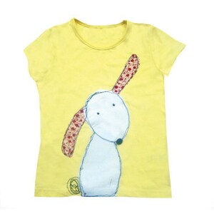 Op de afbeelding: Een geel T-shirt met een witte hondenillustratie. De hond heeft rode en witte oren en een zwarte neus.