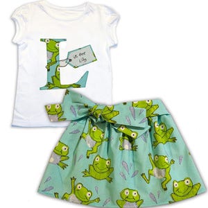 May include: A white short-sleeved t-shirt with a large green letter L on the front. The letter L is made of fabric with a frog pattern. A tag attached to the letter L says "L is for Lily". The t-shirt is paired with a blue skirt with a frog pattern.