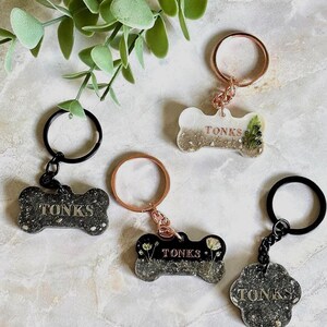 May include: Four keychains in bone and paw shapes, each featuring the name TONKS. Three are black with glitter and one is white with gold glitter. They include rose gold or black metal rings and chains, with embedded dried floral elements.