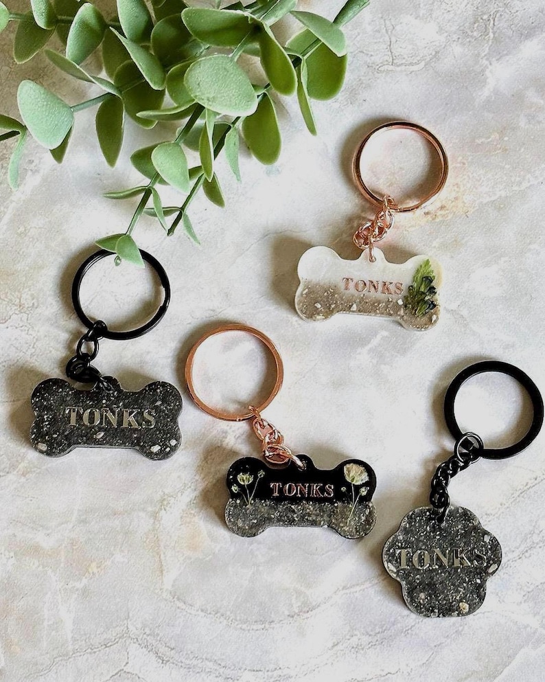 May include: Four keychains in bone and paw shapes, each featuring the name TONKS. Three are black with glitter and one is white with gold glitter. They include rose gold or black metal rings and chains, with embedded dried floral elements.