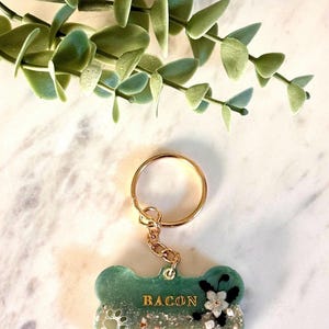 May include: A bone-shaped keychain featuring a green and white resin finish with embedded glitter and sand textures. The name BACON is printed in gold lettering, accompanied by a small white flower and a paw print design attached to a gold ring.