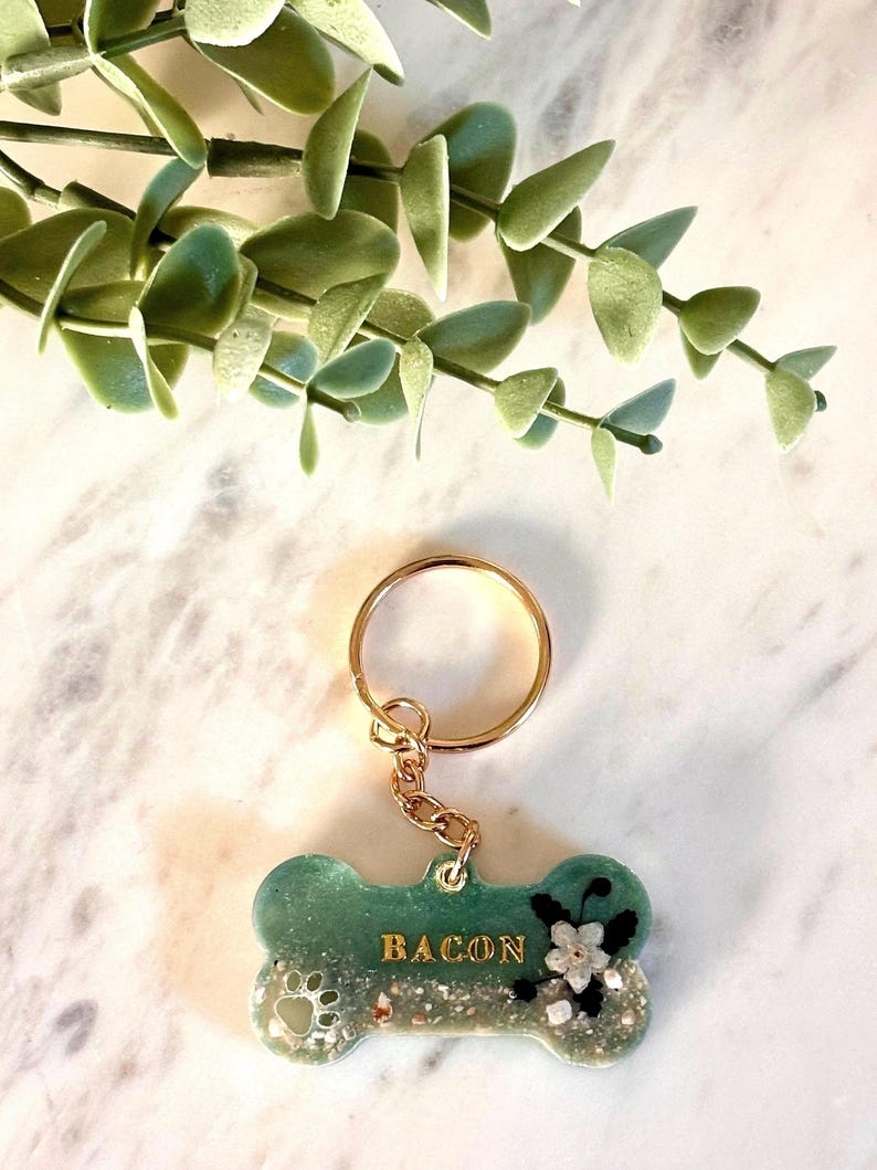 May include: A bone-shaped keychain featuring a green and white resin finish with embedded glitter and sand textures. The name BACON is printed in gold lettering, accompanied by a small white flower and a paw print design attached to a gold ring.