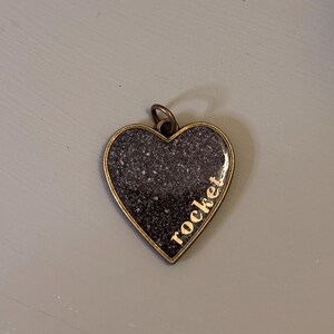 May include: A heart-shaped pendant with an antique bronze-toned metal frame. The center features a dark, speckled, granite-like surface with the word rocket printed in gold-colored lowercase letters along the right side. A small metal jump ring is attached.