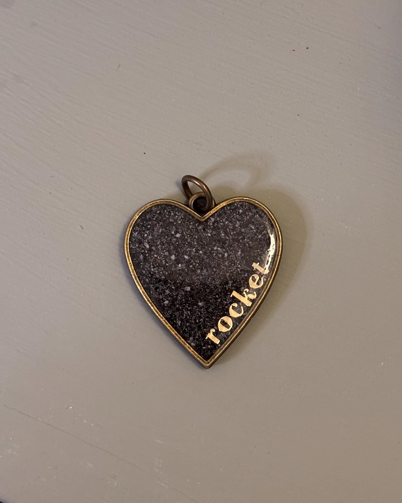 May include: A heart-shaped pendant with an antique bronze-toned metal frame. The center features a dark, speckled, granite-like surface with the word rocket printed in gold-colored lowercase letters along the right side. A small metal jump ring is attached.