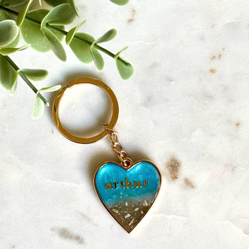 May include: A gold-toned metal keychain featuring a heart-shaped pendant with a resin finish. The pendant displays a blue and white ocean wave design over a sandy base, with the name arthur printed in gold letters across the center.