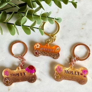 May include: Three bone-shaped keychains made of translucent resin with embedded dried flowers and glitter. Two keychains feature the name MOLLY and one features the name MURPHY in gold lettering. Each includes a metal ring and a small paw print detail.