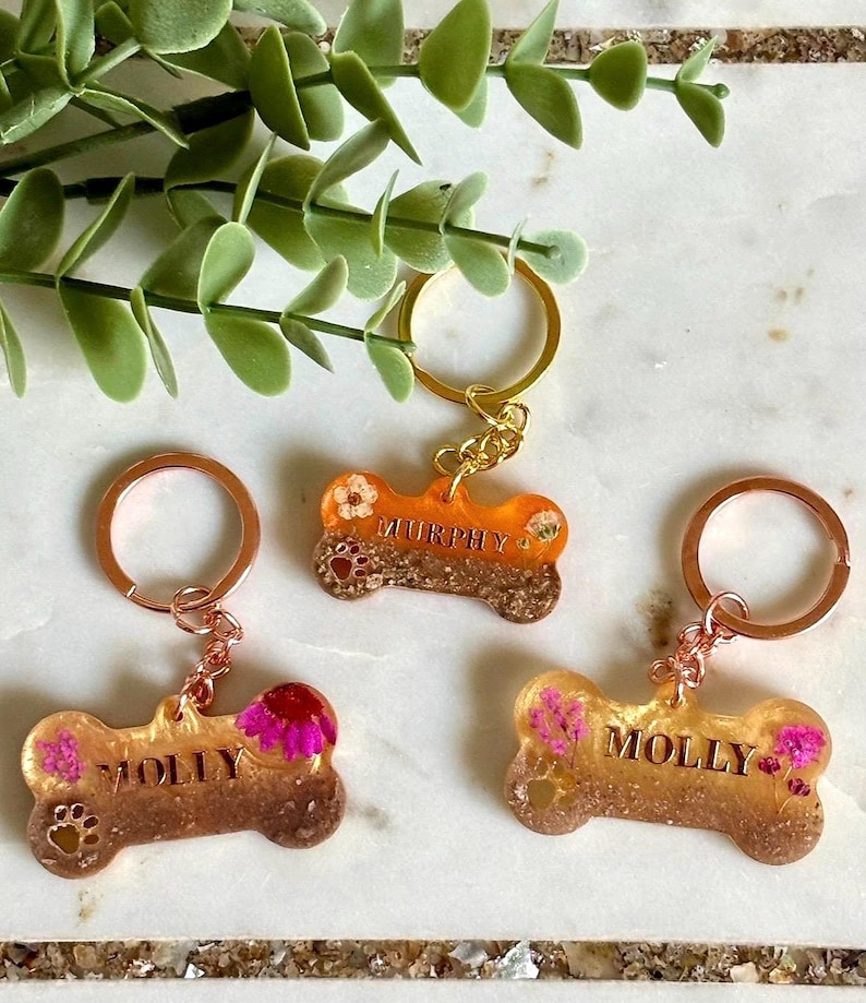 May include: Three bone-shaped keychains made of translucent resin with embedded dried flowers and glitter. Two keychains feature the name MOLLY and one features the name MURPHY in gold lettering. Each includes a metal ring and a small paw print detail.