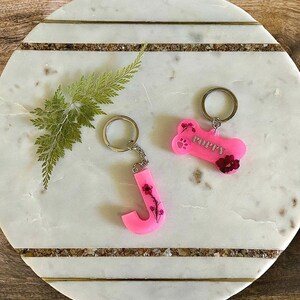 May include: Two pink resin keychains with pressed flowers. One keychain is a bone shape with the word "Poppy" on it. The other keychain is a letter "J".