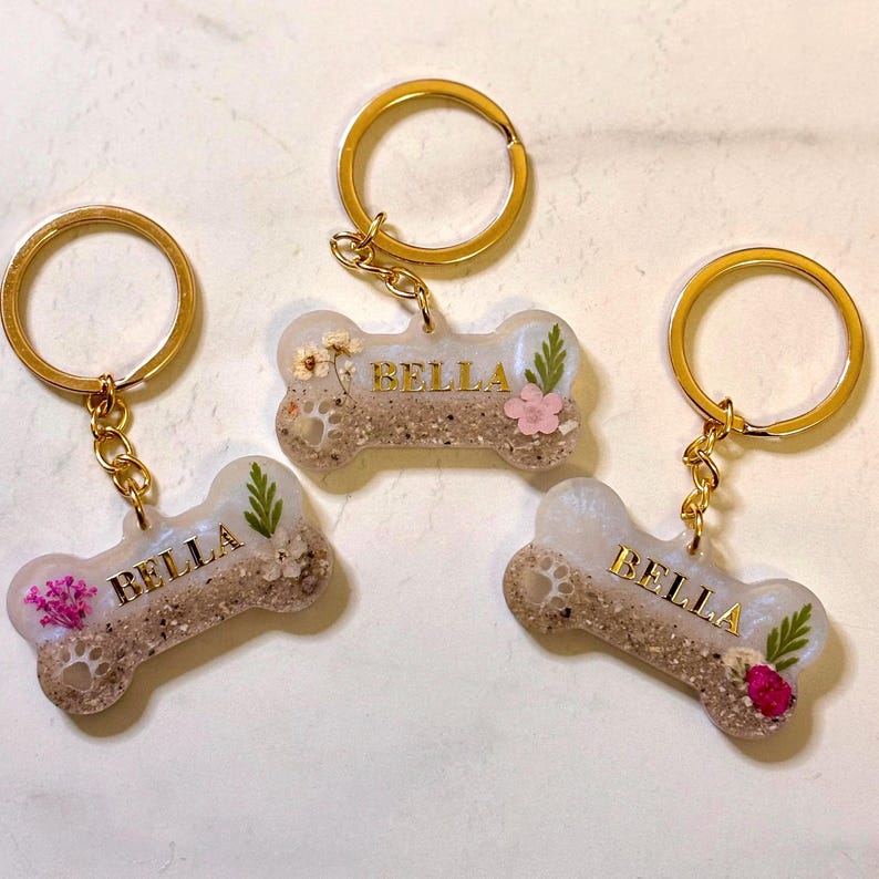 May include: Three bone-shaped keychains with gold-tone metal rings. Each keychain features a clear resin body containing sand, a small paw print impression, embedded dried flowers, and the name BELLA in gold-colored capital letters.