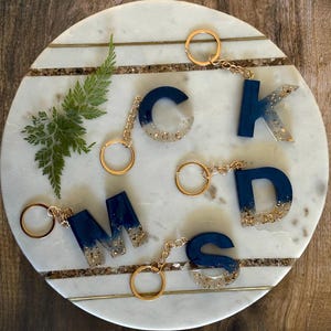 May include: Five resin letter keychains with gold glitter accents. The letters are M, S, D, K, and C. The keychains are on a white marble surface.