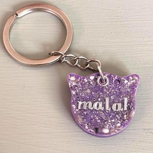 May include: A cat-shaped keychain featuring a purple resin base embedded with small, light-colored flecks and the name "malak" written in silver-toned, lowercase letters. The charm is attached to a circular silver-toned metal split ring.