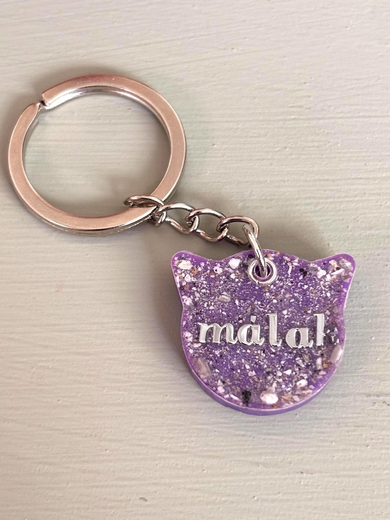 May include: A cat-shaped keychain featuring a purple resin base embedded with small, light-colored flecks and the name "malak" written in silver-toned, lowercase letters. The charm is attached to a circular silver-toned metal split ring.