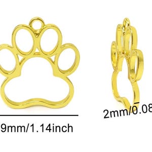 May include: A gold-colored metal paw print charm frame with an open center. The charm measures 32mm (1.26 inches) in height, 29mm (1.14 inches) in width, and has a thickness of 2mm (0.08 inches), featuring a small loop at the top for attachment.