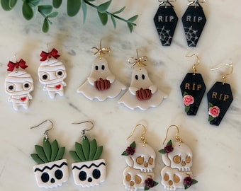May include: A collection of five pairs of earrings featuring Halloween-themed designs including mummies with red bows, ghosts holding pumpkins, black coffins with RIP text, skull planters with green succulents, and stacked skulls with roses.