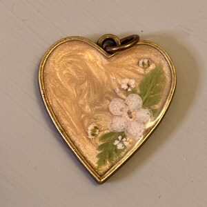 May include: A heart-shaped pendant with a bronze-toned metal frame. The center features a swirling gold-colored resin inlay embedded with small white pressed flowers and delicate green fern leaves. A small circular metal loop is attached at the top.