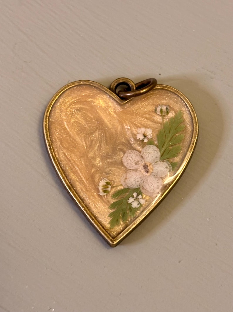 May include: A heart-shaped pendant with a bronze-toned metal frame. The center features a swirling gold-colored resin inlay embedded with small white pressed flowers and delicate green fern leaves. A small circular metal loop is attached at the top.