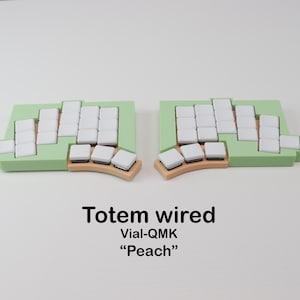 Könnte beinhalten: Eine geteilte Tastatur mit hellgrüner Basis und weißen Tastenkappen. Die Tastatur ist mit "Totem wired Vial-QMK 'Peach'" in schwarzem Text beschriftet. Die Tastatur ist für ergonomische Nutzung konzipiert.