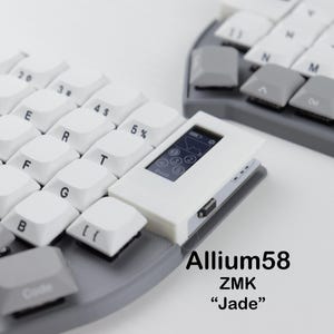 Könnte beinhalten: Eine geteilte ergonomische Tastatur mit weißen Tastenkappen und grauen Akzenten. Ein kleines weißes Gerät mit einem Bildschirm ist angebracht. Die Tastatur ist mit "Allium58 ZMK 'Jade'" beschriftet.