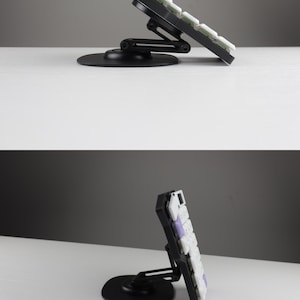 May include: A black mechanical keyboard with white and purple keycaps is mounted on an adjustable black stand. The stand has a circular base and a hinged arm, allowing for tilt adjustment. The keyboard is angled upwards.
