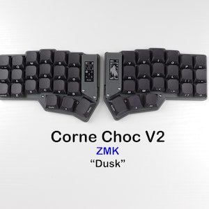 Puede incluir: Un teclado ergonómico dividido con teclas negras, etiquetado como Corne Choc V2 ZMK Dusk. El dispositivo cuenta con dos mitades separadas, cada una con una pequeña pantalla rectangular que muestra iconos y una disposición de teclas escalonada.