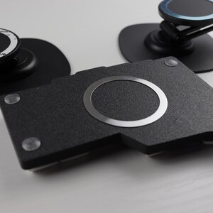 May include: A collection of black phone accessories, including a textured black base with a silver ring, and two phone mounts. The mounts have black bases and adjustable arms. Small, clear adhesive pads are visible.