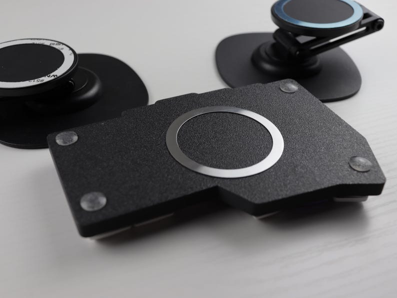 May include: A collection of black phone accessories, including a textured black base with a silver ring, and two phone mounts. The mounts have black bases and adjustable arms. Small, clear adhesive pads are visible.
