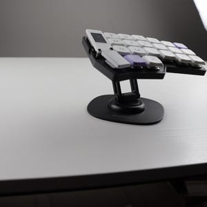 May include: A black ergonomic keyboard with white and purple keycaps. The keyboard is split into two sections and mounted on a black stand. The keys are arranged in a curved layout, designed for comfort and efficiency.