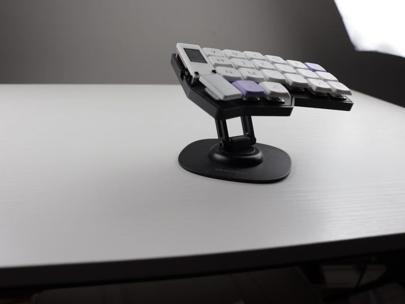 May include: A black ergonomic keyboard with white and purple keycaps. The keyboard is split into two sections and mounted on a black stand. The keys are arranged in a curved layout, designed for comfort and efficiency.