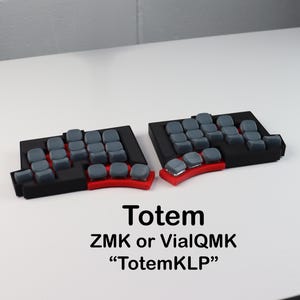 Könnte beinhalten: Zwei schwarze und rote mechanische Tastaturhälften mit grauen Tastenkappen. Die Tastaturen sind mit "Totem" mit dem Text "ZMK or VialQMK" und "TotemKLP" darunter beschriftet. Die Tastaturen befinden sich auf einer weißen Oberfläche.