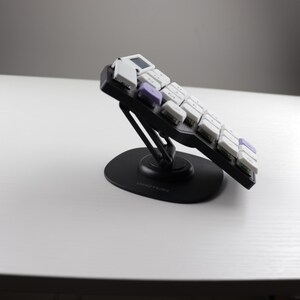 May include: A black ergonomic keyboard with white and purple keys is angled upward on a black stand. The keyboard is on a white surface, with a gray background and a bright light source.